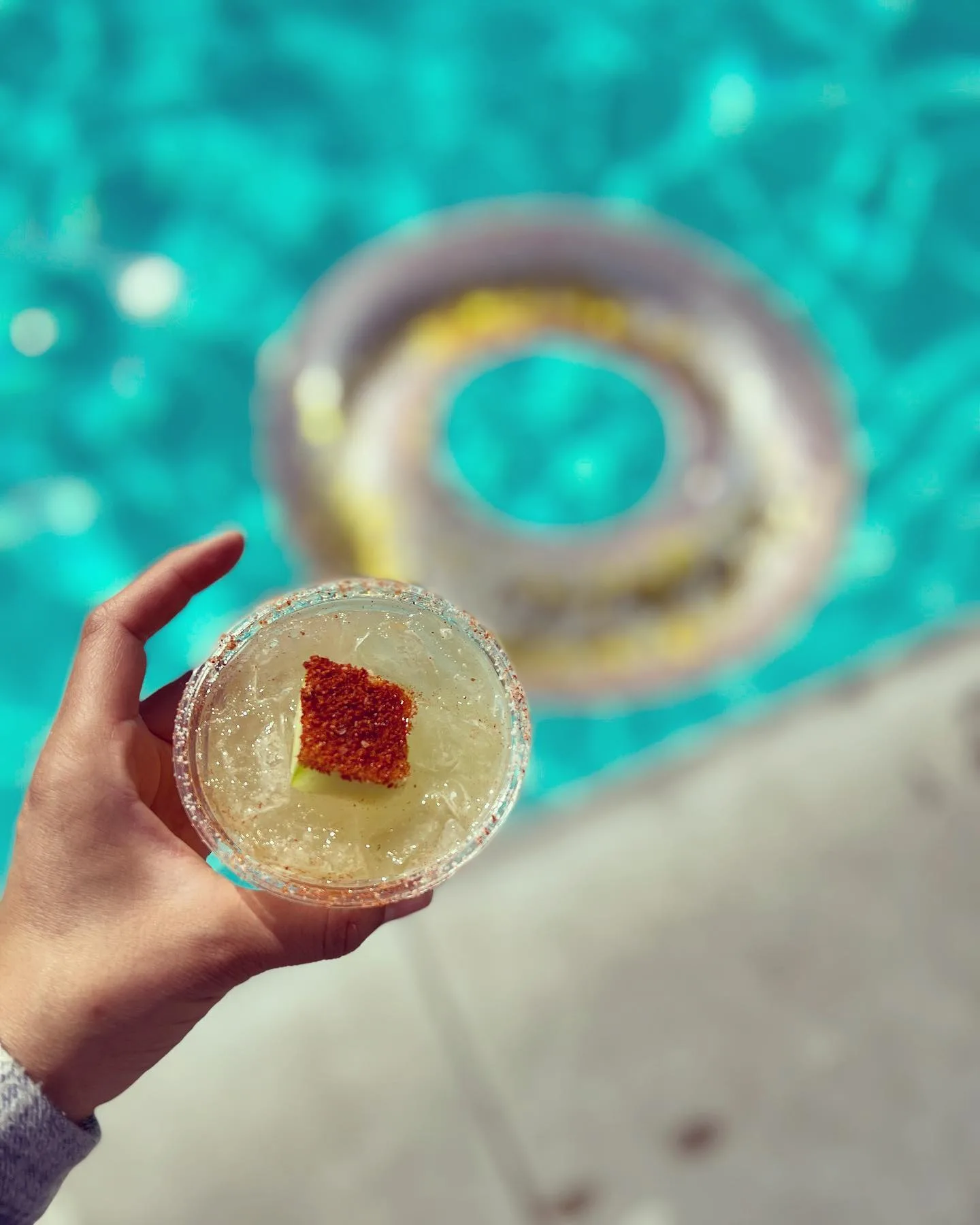Top-down view of a pale garnished cocktail held over turquoise pool water