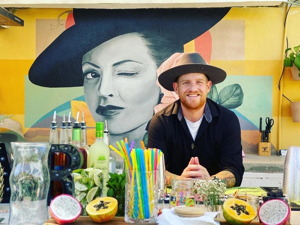 Cameron Alsup smiling behind a colorful cocktail bar